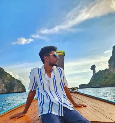 Ashok sitting at the bow of a wooden longtail boat wearing sunglasses and a striped shirt, with limestone karst cliffs and calm blue water behind him